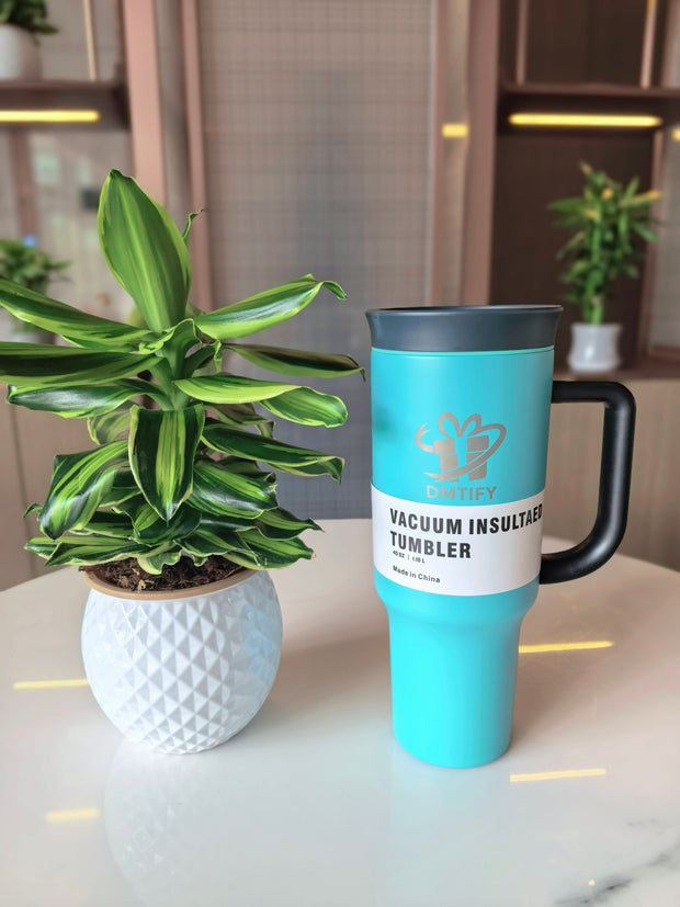Turquoise tumbler with black handle on a surface next to a small potted plant.
