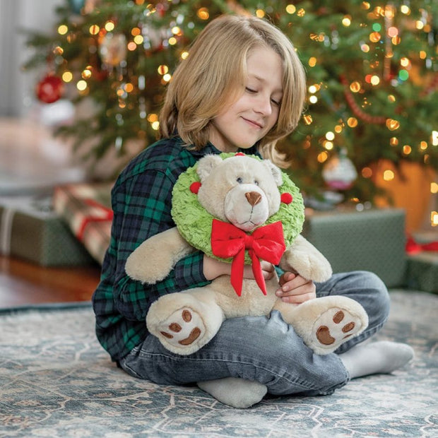 Child holding a teddy bear with a wreath and bow in front of a decorated Christmas tree.