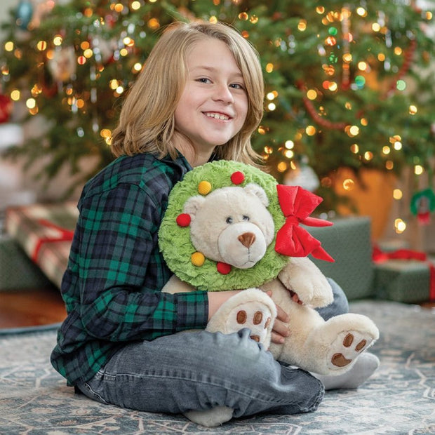 Child holding a teddy bear with a wreath in front of a decorated Christmas tree.