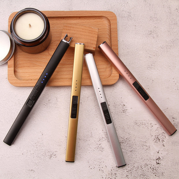 Four candle lighters in black, gold, silver, and rose gold on a wooden tray with candles.