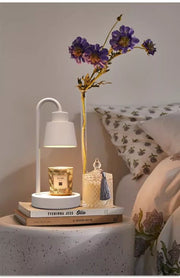 White lamp on a nightstand with books, a small candle, and a vase with purple flowers.