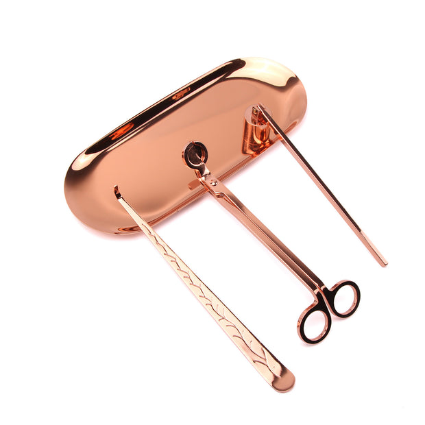 Rose gold Set of candle accessories including wick trimmer, dipper, snuffer and tray on a white surface with decorative items.