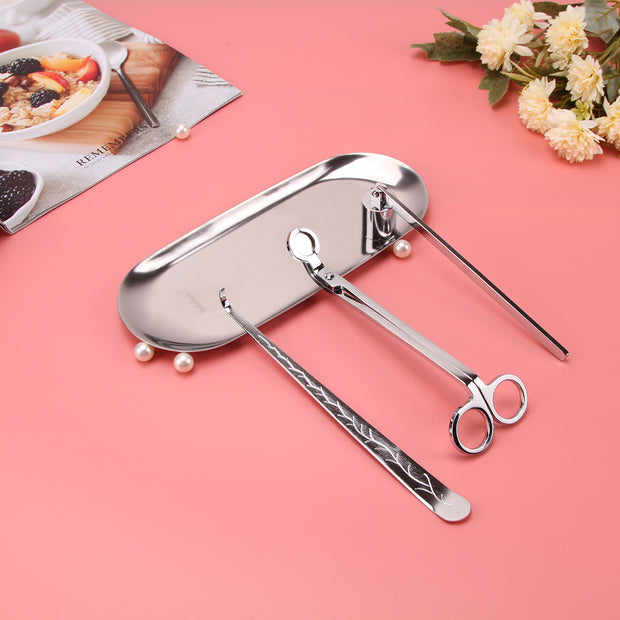 Set of silver candle accessories including wick trimmer, dipper, snuffer and tray on a pink background with a magazine and flowers.