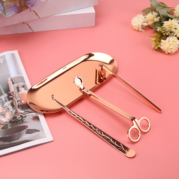 Rose gold Set of candle accessories including wick trimmer, dipper, snuffer and tray on a pink surface with a magazine and flowers in the background.
