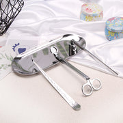 Set of silver candle accessories including wick trimmer, dipper, snuffer and tray on a white surface with decorative items.